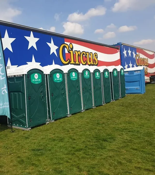 A row of green portable toilets on grass in front of a circus trailer with a red, white, and blue star-spangled design. The sky is clear with a few clouds.