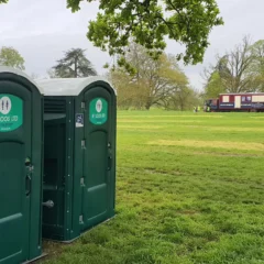 Portable toilets stand on a grassy field under a cloudy sky. A group of shipping containers is visible in the distance. Trees with fresh green leaves frame the scene, suggesting a park or outdoor event setting.