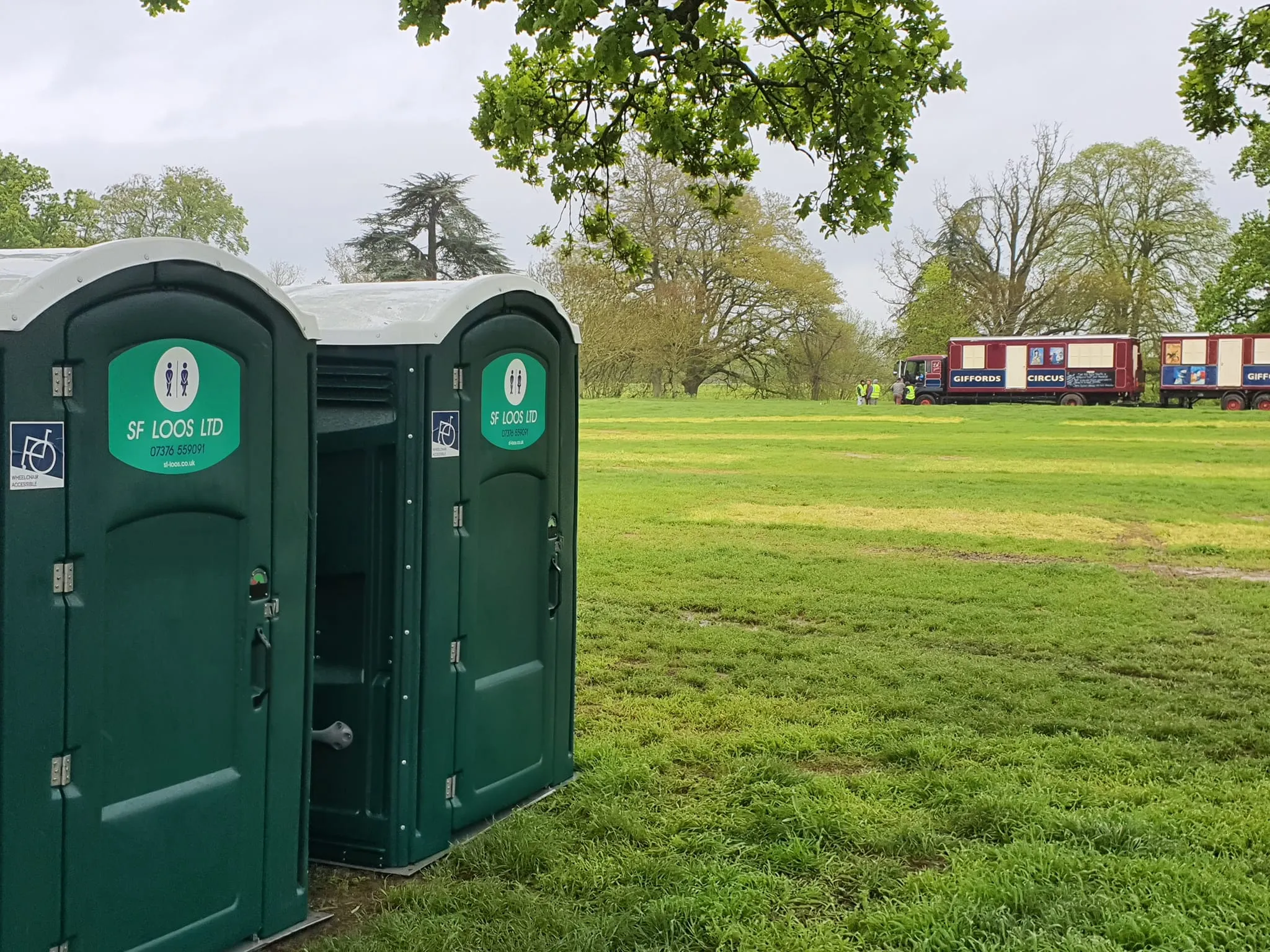 Portable toilets stand on a grassy field under a cloudy sky. A group of shipping containers is visible in the distance. Trees with fresh green leaves frame the scene, suggesting a park or outdoor event setting.