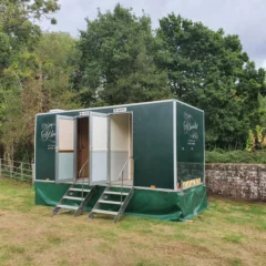 Mobile restroom units situated on grass, surrounded by trees and a stone wall. Two doors labeled "Ladies" and "Gents" with small steps leading up. Overcast sky above.