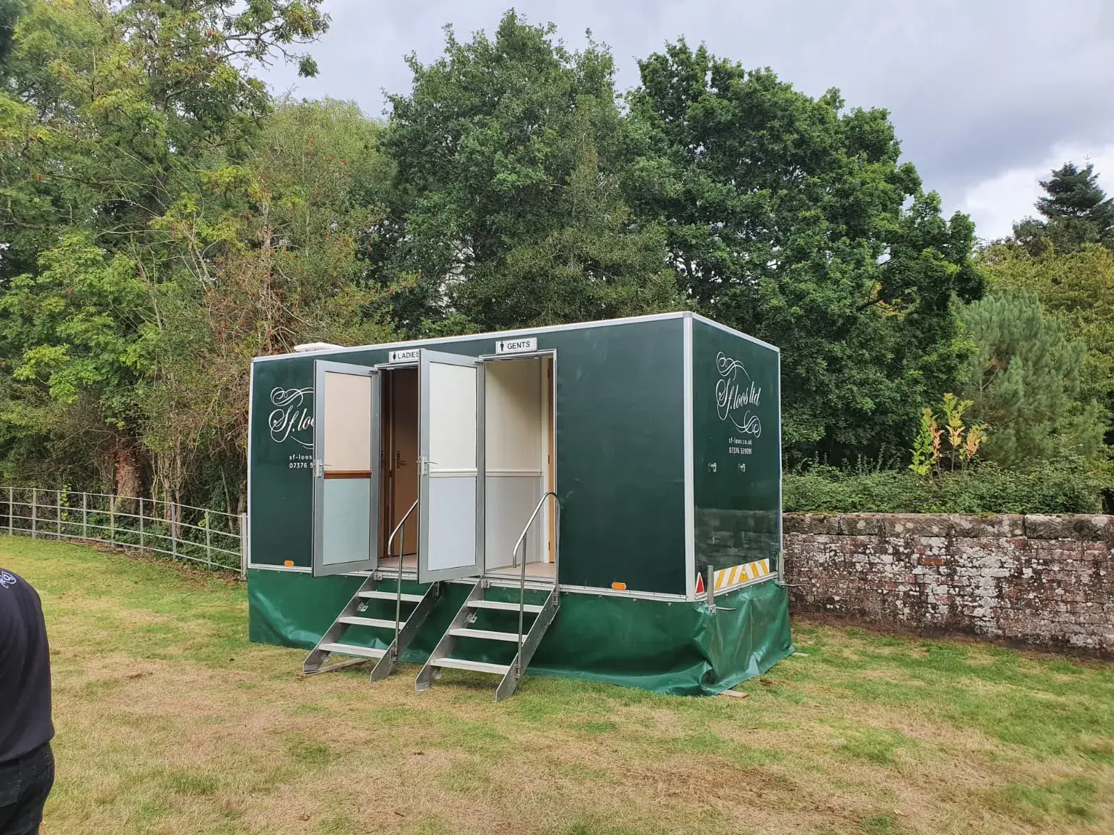 Mobile restroom units situated on grass, surrounded by trees and a stone wall. Two doors labeled "Ladies" and "Gents" with small steps leading up. Overcast sky above.