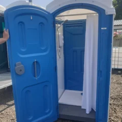 An open blue portable shower unit with a white curtain, situated outdoors on a gravel surface. Sunlit with a clear sky and trees in the background. A hand is visible holding the door open.