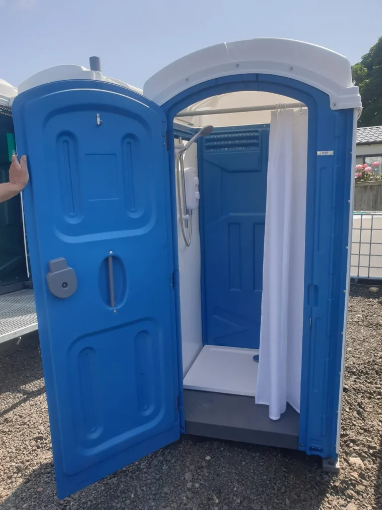 An open blue portable shower unit with a white curtain, situated outdoors on a gravel surface. Sunlit with a clear sky and trees in the background. A hand is visible holding the door open.