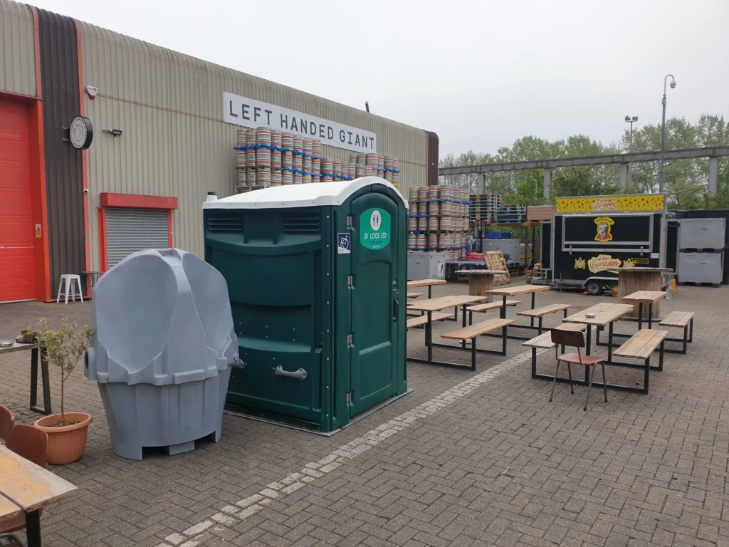 Outdoor seating area with wooden tables and benches. A portable toilet and handwashing station are in the foreground. A food truck is parked nearby, and barrels are stacked against a building labeled "Left Handed Giant." Trees are visible in the background.