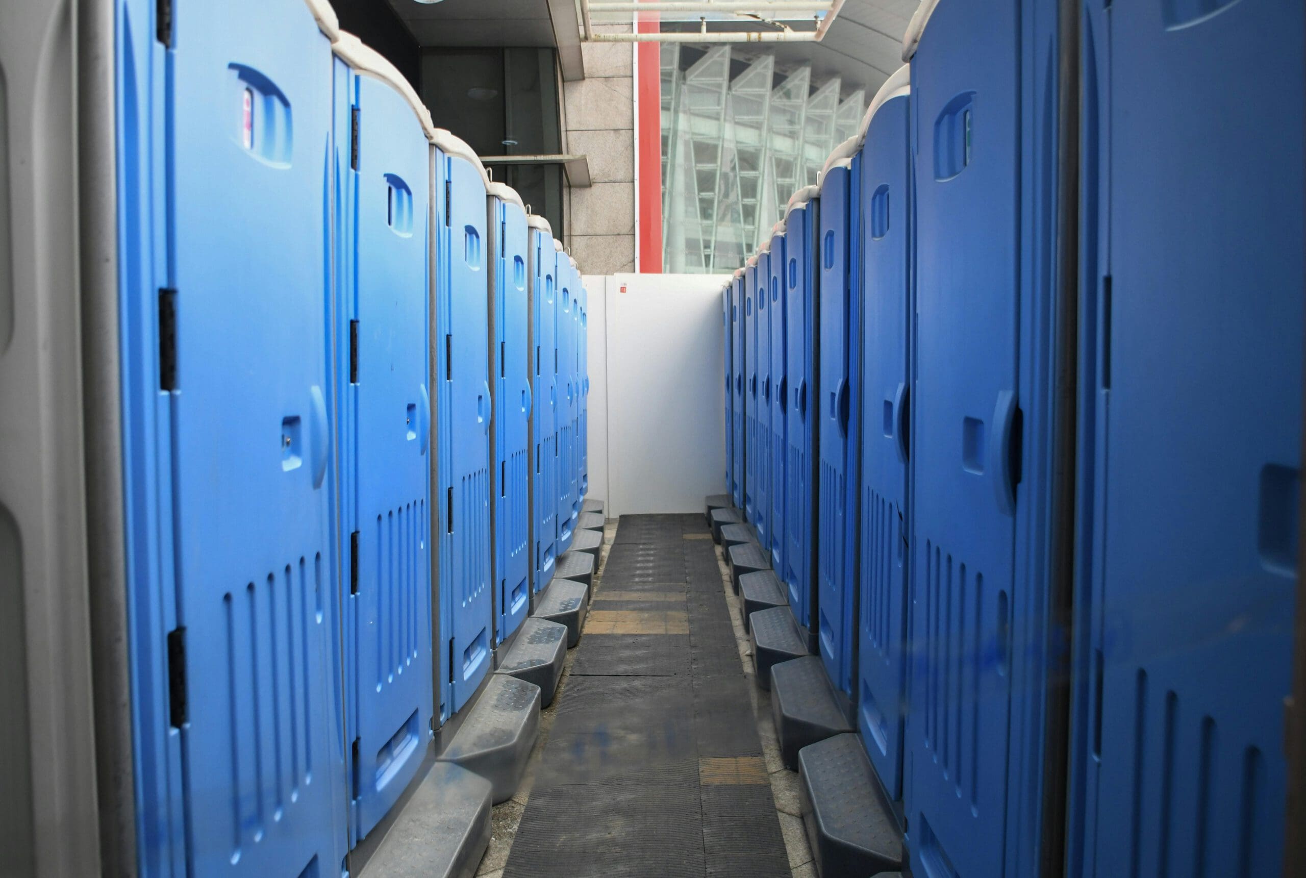 A row of blue portable toilets lined up in an indoor area with a tiled floor and a glass-paneled background. The toilets have doors closed, and there's a narrow walkway between them.