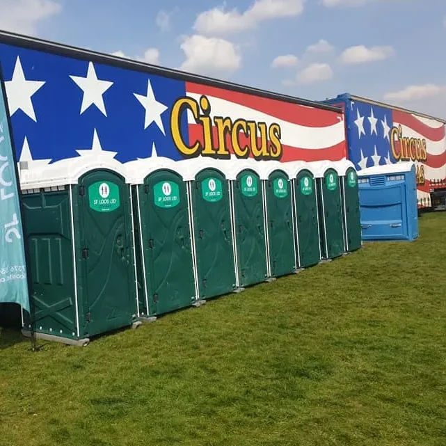 A row of green portable toilets is lined up in front of a large truck with a colorful design featuring stars and the word "Circus" in bold letters. The scene is set on a grassy field under a partly cloudy sky.