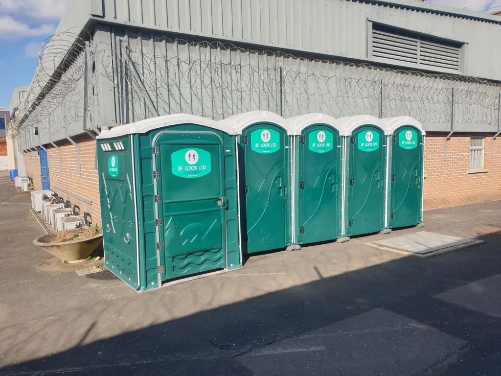 Four green portable toilets are lined up against a brick and metal building with barbed wire on the roof. The area is sunny with a clear sky.