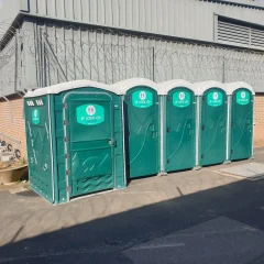 Four green portable toilets are lined up against a brick and metal building with barbed wire on the roof. The area is sunny with a clear sky.